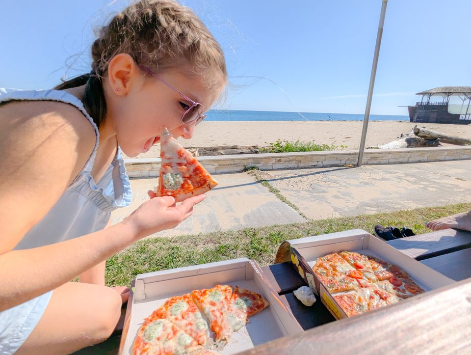 Kind isst Pizza aus dem Karton auf einer Bank am Strand von Obzor
