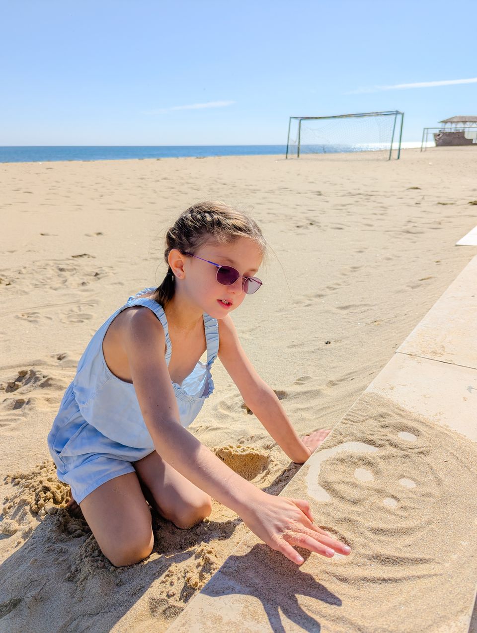 Mädchen malt am Strand ein Bild in den Sand, im Hintergrund Fußballtore